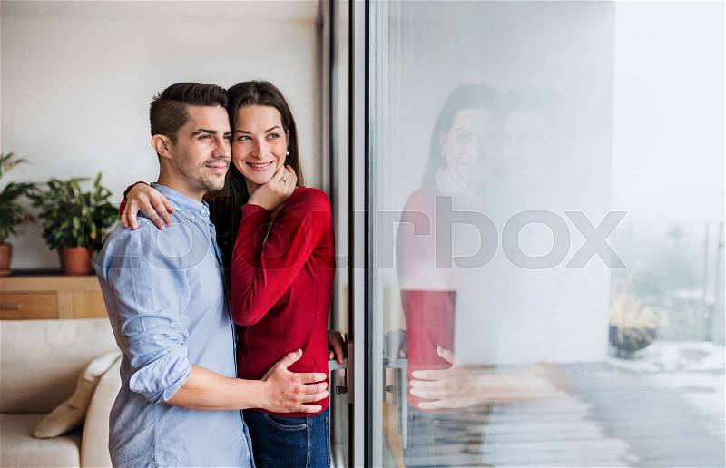 Young happy couple in love standing by the window at home, hugging. Copy space, stock photo