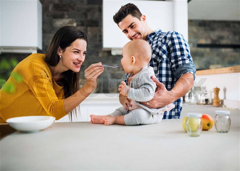 A portrait of young family standing in a kitchen at home, a man holding a baby and a woman feeding her with a spoon, stock photo