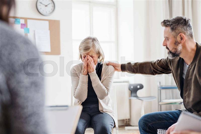 A senior depressed woman crying during group therapy, other people comforting her, stock photo