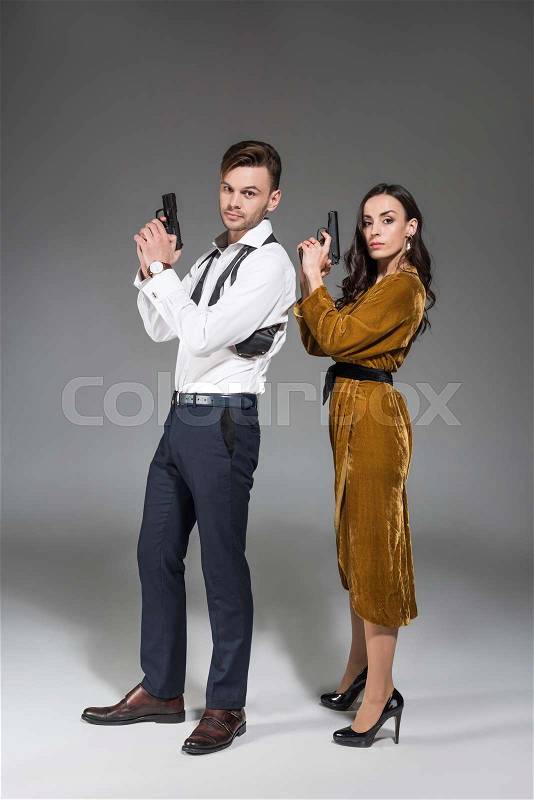 Beautiful confident couple of secret agents posing with gun on grey, stock photo