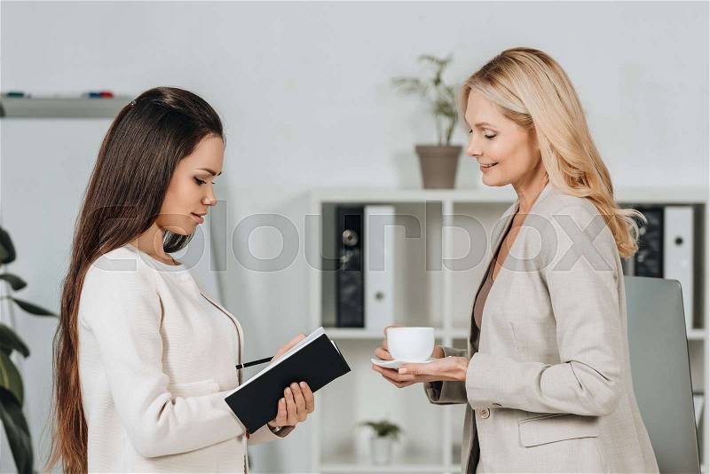 Side view of smiling business mentor with cup of coffee looking at young female colleague taking notes in notebook, stock photo