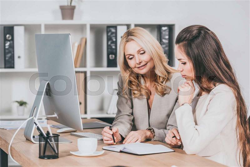 Smiling female business mentor pointing at notebook and working with young colleague in office, stock photo