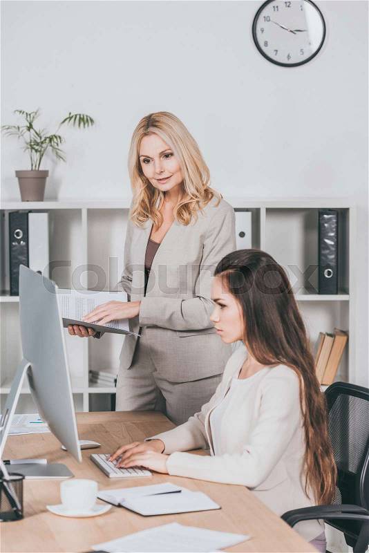 Mature business mentor with folder and young businesswoman looking at desktop computer in office, stock photo
