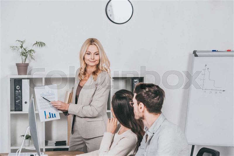 Smiling business mentor pointing at charts and looking at young colleagues sitting in office, stock photo