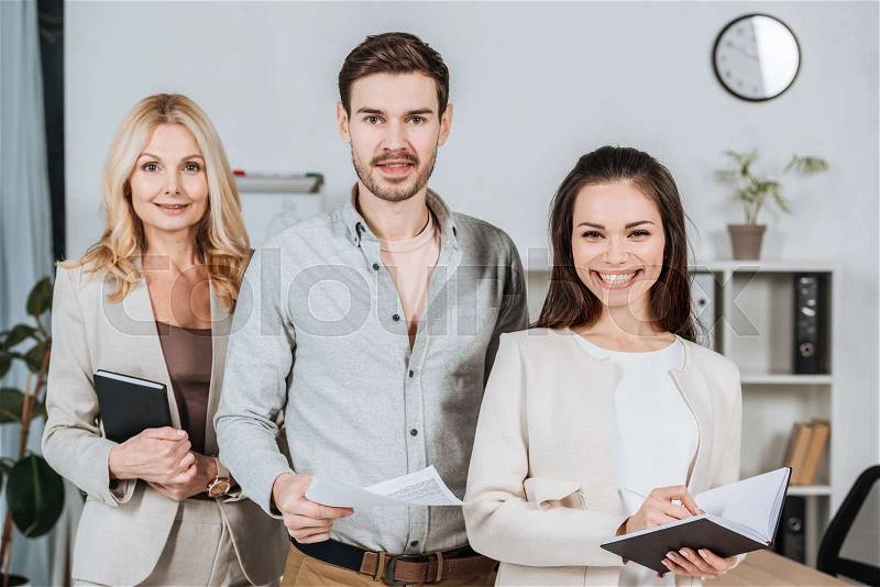 Professional mature business mentor with young colleagues standing together and smiling at camera in office, stock photo