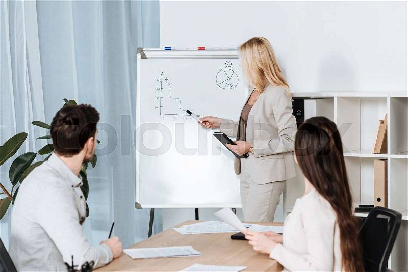 Business mentor pointing at whiteboard with charts while young colleagues sitting at table in office, stock photo