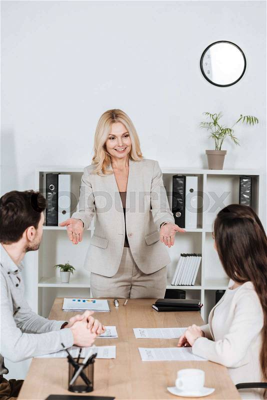 Smiling female business mentor looking at young colleagues sitting at workplace, stock photo