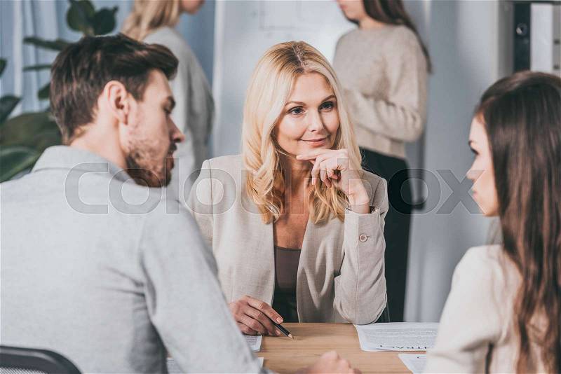 Beautiful smiling business mentor sitting with hand on chin and looking at young colleagues in office, stock photo