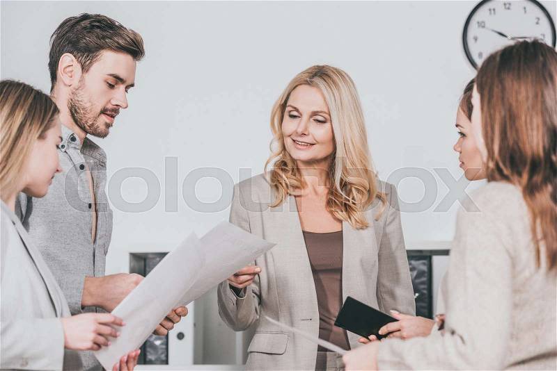 Smiling professional business mentor looking at papers and working with young colleagues in office, stock photo