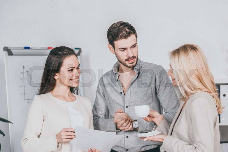 Smiling business mentor holding cup of coffee and talking with young colleagues in office, stock photo