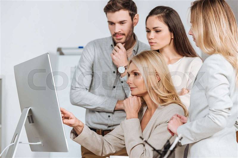 Mature business mentor pointing at desktop computer and working with young colleagues in office, stock photo