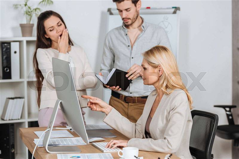 Professional business mentor pointing at desktop computer and working with bored young colleagues in office, stock photo