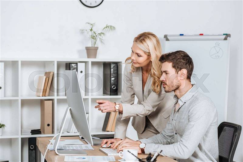 Professional business mentor pointing at desktop computer and working with young colleague in office, stock photo