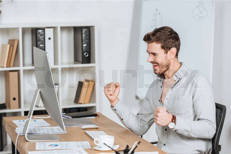 Excited young businessman shaking fists and looking at desktop computer in office, stock photo