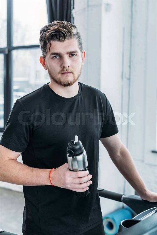 Handsome young man holding sports bottle and looking at camera in gym, stock photo