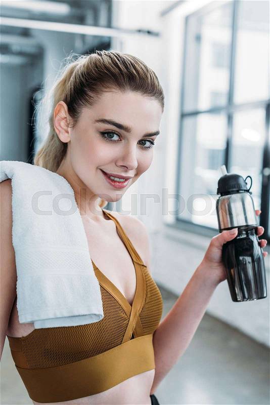 Attractive sporty girl with towel on shoulder holding sports bottle and smiling at camera in gym, stock photo