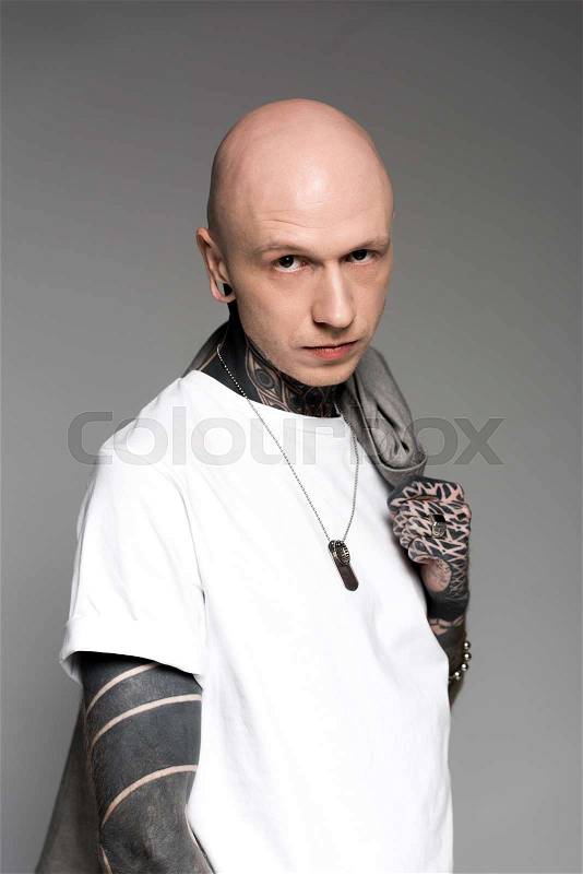 Serious tattooed man in white t-shirt holding suit jacket on shoulder and looking at camera isolated on grey, stock photo