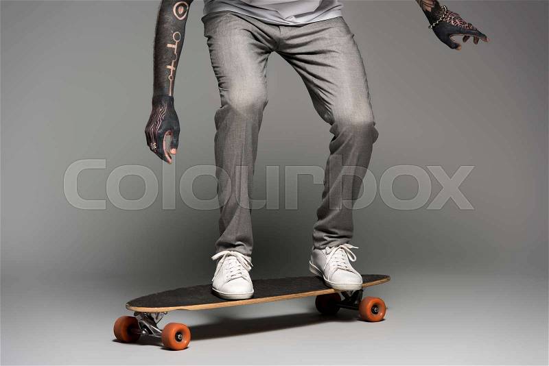 Low section of stylish tattooed man balancing on skateboard on grey, stock photo