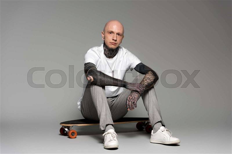 Bald tattooed man sitting on skateboard and looking at camera on grey, stock photo