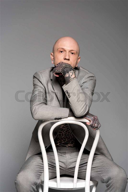 Thoughtful tattooed man in suit sitting with hand on chin and looking at camera isolated on grey, stock photo