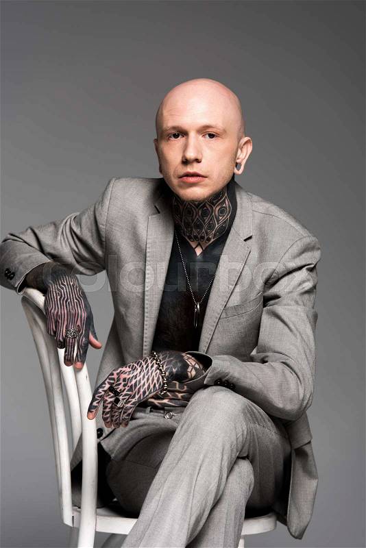 Confident bald tattooed man in suit sitting on chair and looking at camera isolated on grey , stock photo