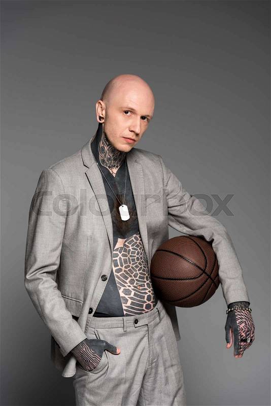 Stylish bald tattooed man in suit holding basketball ball and looking at camera isolated on grey, stock photo