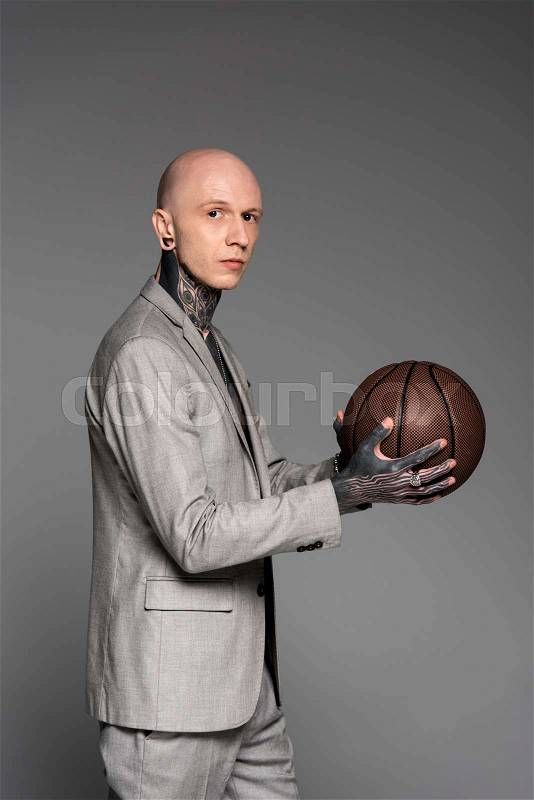 Side view of bald tattooed man in suit holding basketball ball and looking at camera isolated on grey, stock photo