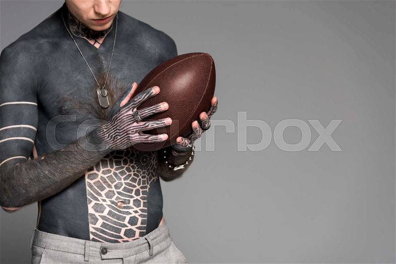 Cropped shot of bare-chested man with tattoos holding rugby ball isolated on grey, stock photo