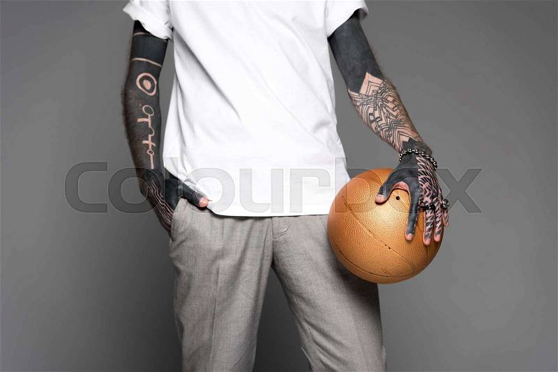 Cropped shot of tattooed man in white t-shirt standing with golden basketball ball isolated on grey, stock photo