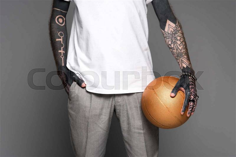 Cropped shot of tattooed man standing with hand in pocket and holding golden basketball ball isolated on grey, stock photo