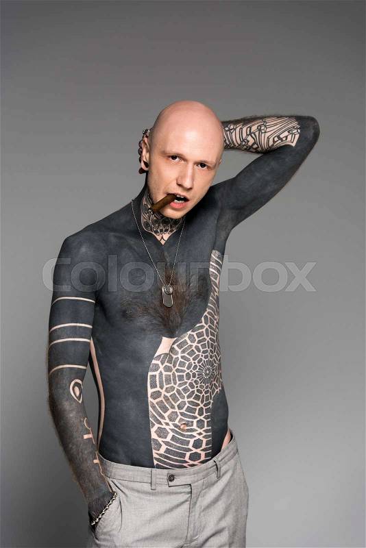 Bald shirtless tattooed man with hand behind head smoking cigar and looking at camera isolated on grey, stock photo