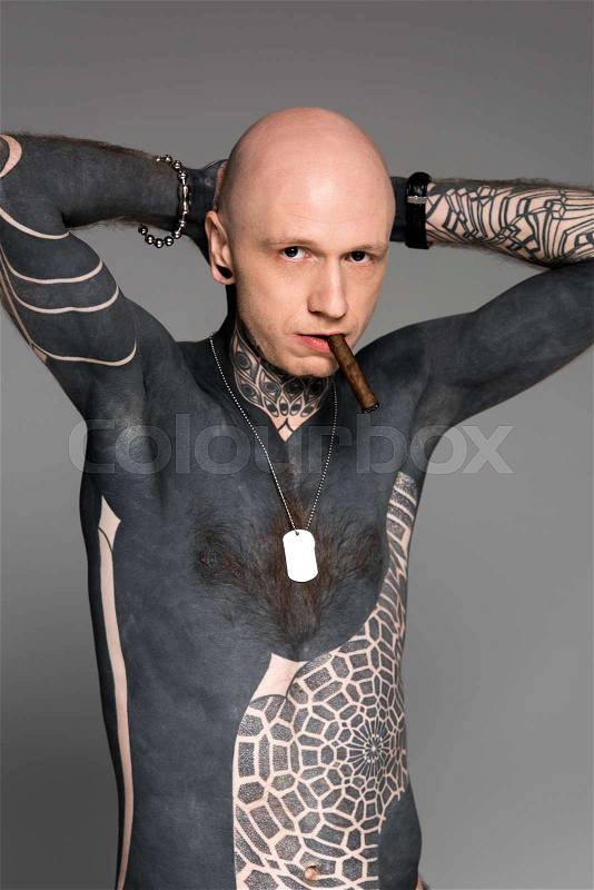 Confident tattooed bare-chested man with hands behind head smoking cigar and looking at camera isolated on grey, stock photo