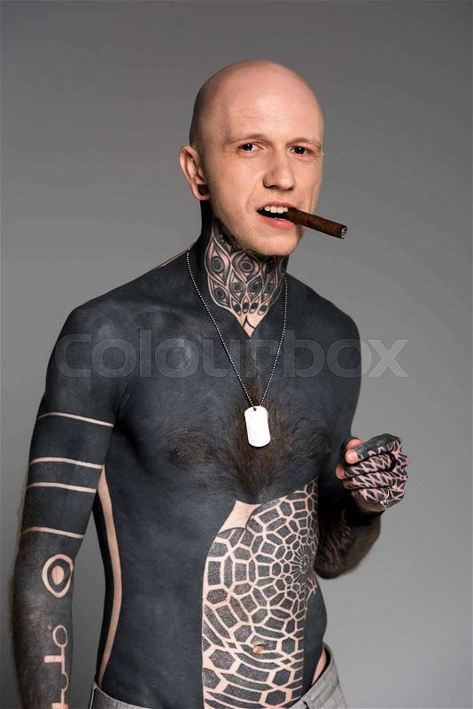 Confident bare-chested man with tattoos smoking cigar and looking at camera isolated on grey, stock photo