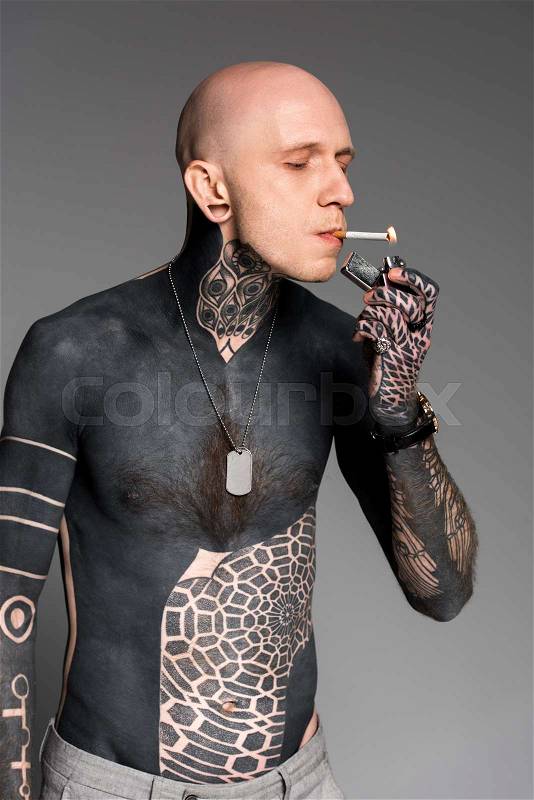 Bald shirtless tattooed man lighting cigarette with lighter isolated on grey, stock photo