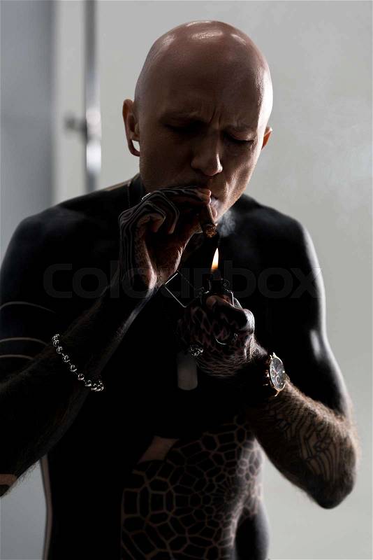 Bare-chested man with tattoos lighting cigar with lighter on grey, stock photo