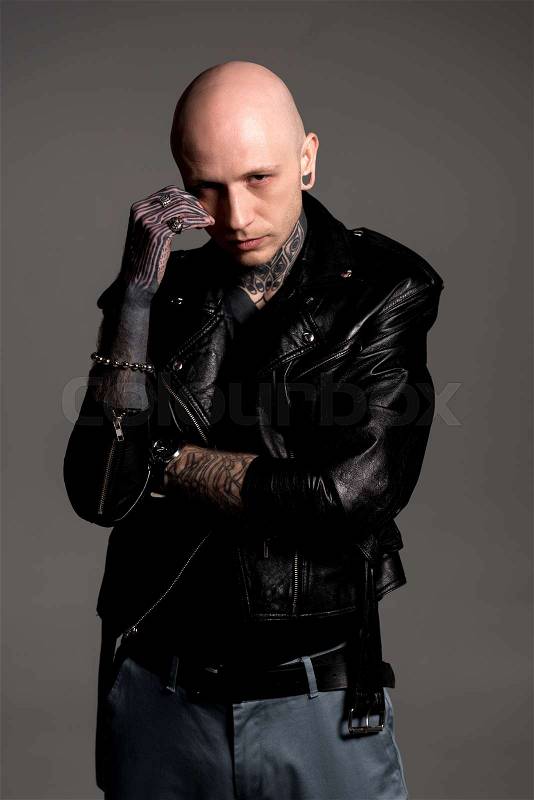 Confident tattooed man in leather jacket standing with hand on face and looking at camera isolated on grey , stock photo