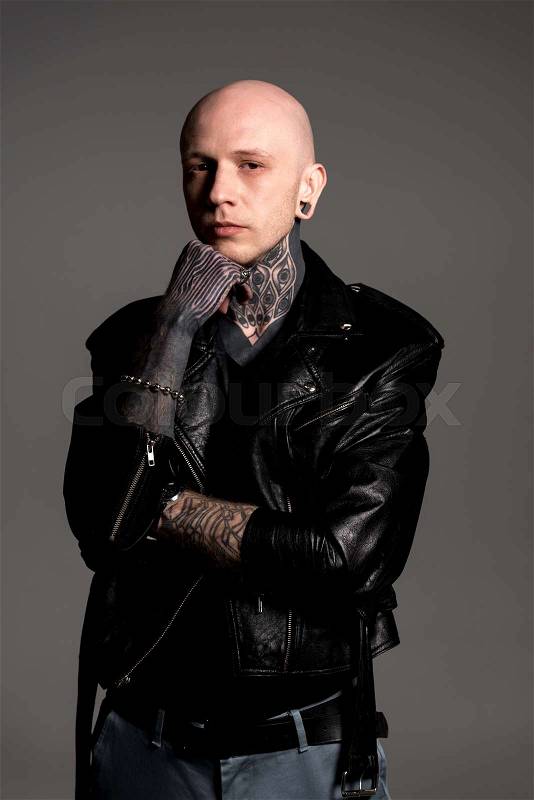 Confident tattooed man in leather jacket standing with hand on chin and looking at camera isolated on grey , stock photo