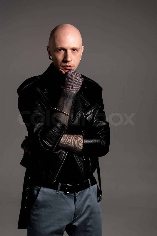 Stylish tattooed man in leather jacket standing with hand on chin and looking at camera isolated on grey, stock photo