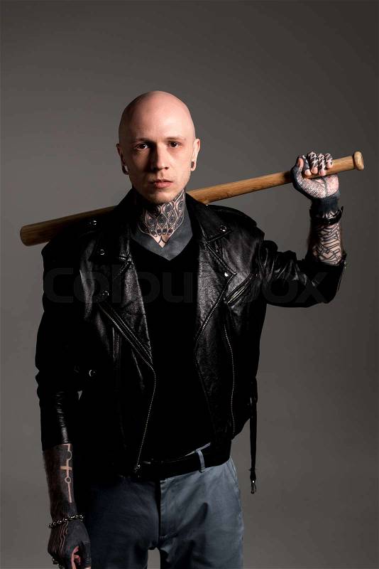 Cool tattooed man in leather jacket holding baseball bat on shoulders and looking at camera isolated on grey, stock photo