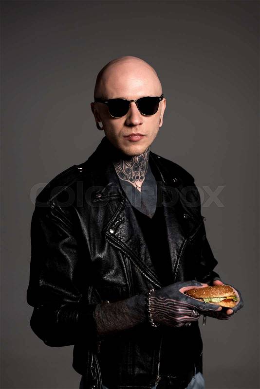 Tattooed man in leather jacket and sunglasses holding tasty burger and looking at camera isolated on grey, stock photo