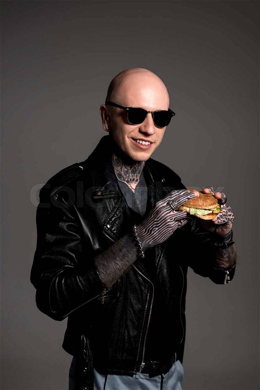 Tattooed man in leather jacket and sunglasses holding tasty burger and smiling at camera isolated on grey , stock photo