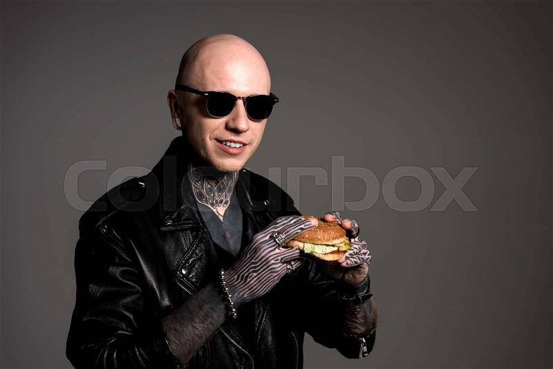 Handsome tattooed man in leather jacket and sunglasses holding hamburger and smiling at camera isolated on grey, stock photo