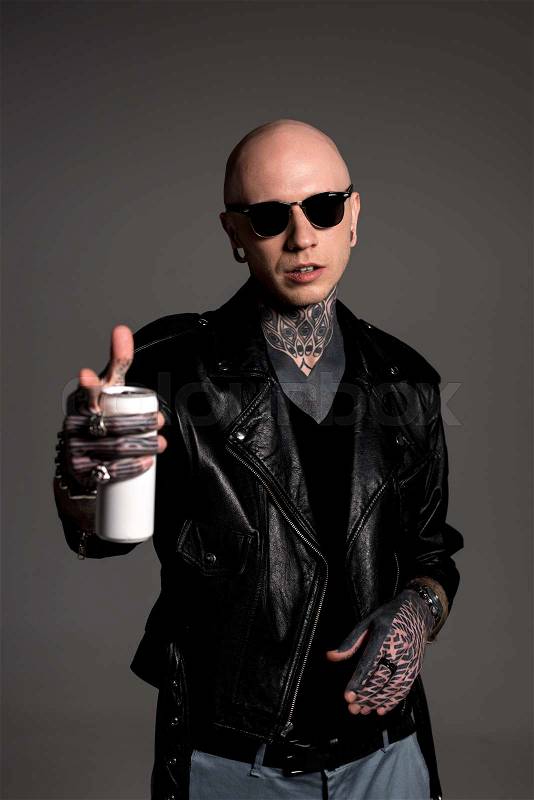 Bald tattooed man in leather jacket and sunglasses holding soda can and pointing at camera isolated on grey, stock photo