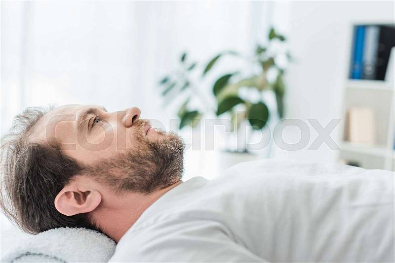 Side view of bearded man lying on massage table and looking up in medical office, stock photo