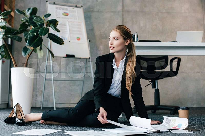 Barefoot beautiful businesswoman sitting on floor near work-table and looking away, stock photo
