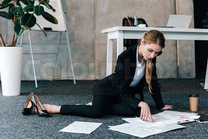 Barefoot businesswoman sitting on floor near work-table and reading documents, stock photo