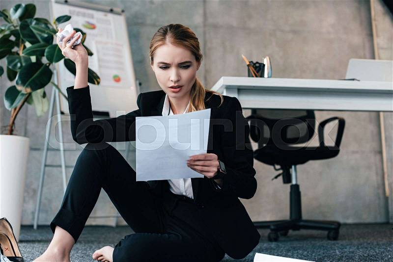 Barefoot businesswoman sitting on floor near work-table with crumpled paper and reading documents, stock photo