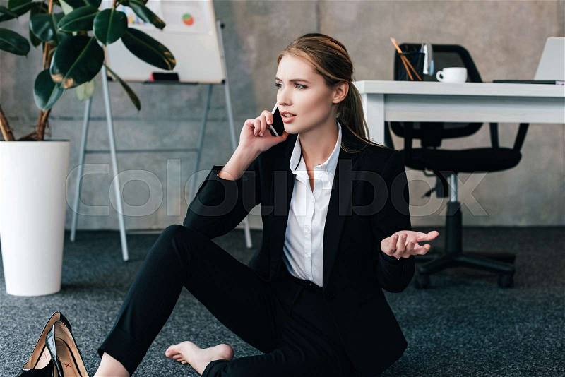 Barefoot businesswoman sitting on floor and talking on smartphone with emotion, stock photo