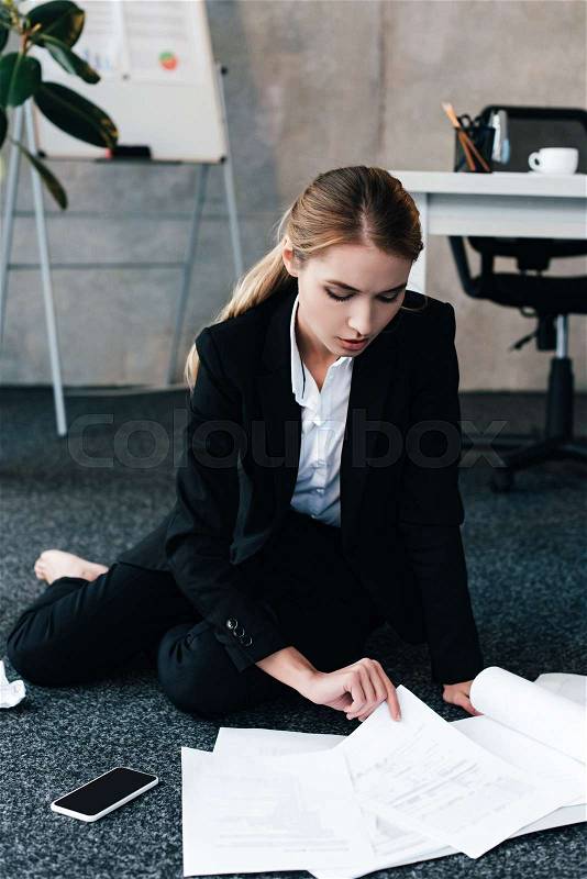 Barefoot businesswoman sitting on floor near work-table and reading documents, stock photo