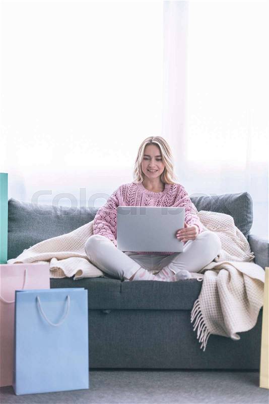 Happy blonde woman sitting on sofa with laptop near shopping bags, stock photo